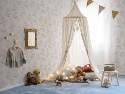 Et barnerom tapetsert med Lycke Vildrosor 1. Gulvet har et blått teppe og det henger en hvit sengehimmel fra taket med puter og bamser inni på gulvet. Speil og en søt kjole henger på veggen. Girlander med farger i taket.