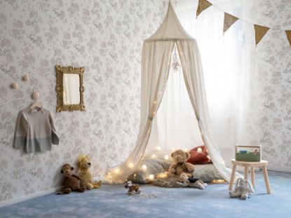 Et barnerom tapetsert med Lycke Vildrosor 2. Gulvet har et blått teppe og det henger en hvit sengehimmel fra taket med puter og bamser inni på gulvet. Speil og en søt kjole henger på veggen. Girlander med farger i taket.