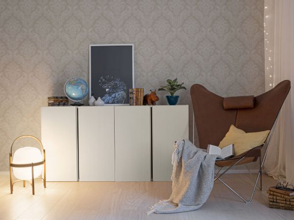 Lycke Historia 1 på vegg i en stue. Inntil veggen står et hvit skap, med dekor av bilde, globus, plante og annet nips oppå. Det står en brun skinnstol foran, med et grått teppe og gul puti oppi. En liten lampe står også på gulvet ved siden av skapet.