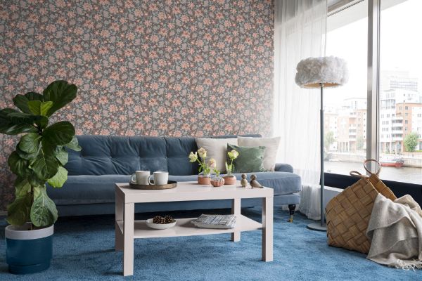 Lycke Nypros 5 tapetsert på en stuevegg bak en blå veloursofa. Gulvteppe er i samme fargetone som sofaen. Det er et stort vindu til høyre i rommet med utsikt mot en elv. Det er hvite detaljer både i form av lette tynne gardiner, en hvit skyformet lampe og et hvitt stuebord fra Ikea. Rommet er myket opp med en bambuskurv og en stor grønnplante, samt grønne og beige puter i sofaen og noe nips i naturfarge på bordet.