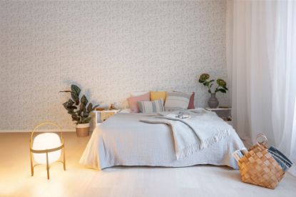Lycke Primula 1 på soveromsvegg bak en seng med hvitt sengetøy og gule og rosa puter. Det er grønnplanter på hver side av sengen, og en lampe på gulvet. En søt naturkurv er også plassert foran sengen. Det henger hvit gardiner foran vinduene ved siden av sengen.